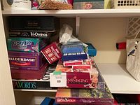 Stack of boxed board games on shelf with visible game titles Boggle Master, Balderdash, Master Craniums, Upwords, Reminiscing, Monopoly Napa Valley Edition.