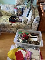 Plastic bins filled with Christmas decorations, variety of hanging ornaments, wrapped paper and tissue on a couch and floor