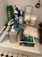 Photo of assorted cleaning supplies arranged in a cabinet including Vim, Lysol, vinyl gloves box, drain stoppers, soap pump bottle, paper towel roll, and cleaning accessories.