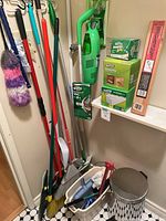 Overall view showing brooms, mops, dusters, Swiffer Sweeper Vac items, Woodpegger rack on shelf, and stainless steel garbage can on floor.