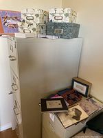 Four decorative photo storage boxes stacked, three floral pattern and one leafy green pattern, placed on a filing cabinet and a white table with additional books and notebooks nearby.