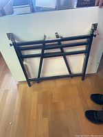 Photo of the folding black metal frame of the drafting table in folded position, showing structural bars and hinges.