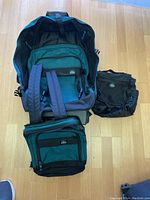Three Eagle Creek travel bags on wooden floor: large wheeled carry-on with backpack straps, small backpack, and a toiletries bag.