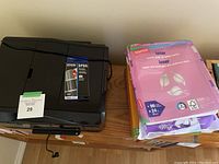 Epson XP-810 printer on a wooden surface with ink cartridge packaging and several packs of printer paper next to it.