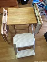 Three wooden folding tables shown fully folded and standing with one table open on wooden floor with light finish. One white Rubbermaid 2-step plastic stool is in front.