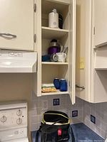 Kitchen cabinet with purple hand blender, white coffee grinder, glass carafe, blue water bottle inside.