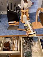 Kitchen counter showing knife block with knives, utensil holder with white plastic utensils, wooden paper towel holder base, meat cleaver, knife sharpener, and open drawers with cutlery and cooking accessories.
