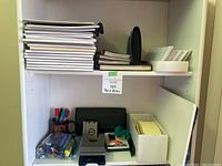 Shelf with multiple stacked notepads, clear pouch of multi-colored markers, black 2-hole punch, sticky notes organizer, and white envelopes.