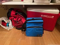 Red Cross emergency backpack next to blue insulated lunch kit and large red and white Coleman cooler on floor.
