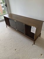 Side angled view of the wooden stereo cabinet showing its overall shape, glass doors, and wood slatted side panels.