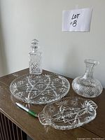 Overall view of five crystal glassware pieces arranged on a wooden surface, including a Scotch decanter, wine vase, large round platter, small platter, and pickle dish with a pen for scale.