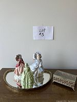 Front and top view of three porcelain figurines, ornate mirror tray, and decorative jewellery box.