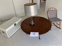 Photo of white wicker chest, round wooden coffee table with a silver table lamp, and antique rocking chair with cushion in background.