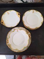 Photo showing the monogrammed white porcelain plates with scalloped gold and green edges, two sizes visible and shallow dish with handles.