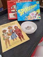 Lot contents showing two Spirograph boxes, the Wedgwood ceramic dish, and Shirley Temple paper dolls book arranged on a black surface.
