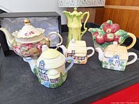 Group photo showing six teapots on table including three miniature cottage teapots, one floral Victorian style teapot, one art nouveau style coffee pot, one apple basket teapot
