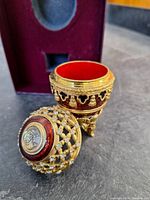 Gold-tone decorative egg with clear crystals and red enamel, hinged open showing red interior, velvet-lined box in background