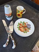 Photo of the ceramic vase with sunset design, sunflower mug, fruit plates, and floral-handled serving utensils arranged on table surface.