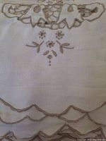 Close-up of scalloped lace edge and floral embroidery on the linen fabric, showing cut lace detail in beige or tan thread on white fabric.