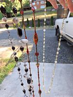 Long colorful bead necklaces held up to light outdoors showing various bead styles and colors including black, white, orange, and faux pearl beads.