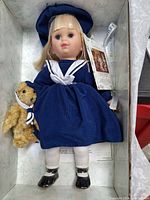 Blonde doll in navy sailor dress with matching hat, black shoes, and white tights showing some stains on back, holding a teddy bear with scarf, all items placed in an open box.
