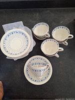 Photo of 4 teacups and 2 saucers, one saucer on top of plastic container, showing blue floral pattern on white ceramic, arranged on black surface