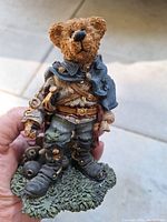 Front view of Boyd Bear figurine in detailed soldier costume standing on grass-textured base with various accessories and a sword.
