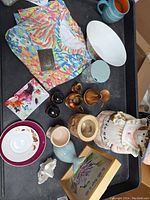 Photo showing 16 miscellaneous resale items on a black tray including clothing, ceramics, glassware, and decorative pieces.