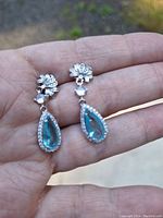 Close-up photo of a pair of sterling silver drop earrings held in hand, featuring blue teardrop-shaped topaz stones surrounded by small clear stones, with a floral stud design.