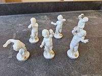 Overview of six small white porcelain cherub figurines with gold detailing on their bases and hands, shown from top front angle on a dark surface.