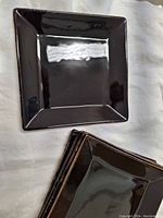 Single square black salad plate with glossy reflective finish and textured rim, stacked plates visible.