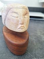 Front view of stone head sculpture on wooden base showing carved facial features and texture of hair.