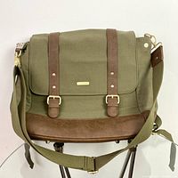 Front view of green canvas backpack with brown faux leather bottom and straps, showing flap and adjustable shoulder strap.