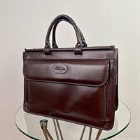 Front view of vintage brown leather briefcase showing handles, embossed patch on front flap pocket, and overall condition.