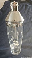 Front view of cocktail shaker showing glass body with white atomic starburst pattern and chrome lid.