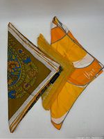 Three scarves displayed, two large square silk scarves with gold and floral paisley patterns, and one smaller synthetic mix scarf in orange tones.