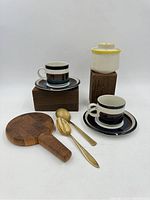 Full view of all items in lot showing two cups and saucers, teak cheeseboard, brass spoons, sugar bowl on elevated wood blocks.