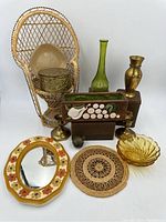 Photo showing entire lot with wicker peacock style plant stand chair in background, amber glass bowl, brass candlesticks, green glass vase, brass vase, floral metal box, brown floral planter, woven trivet, and yellow ochre floral wall mirror.