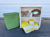 White wicker shelf with Syroco butterfly and mirror on top shelf, two Avon bottles, and folded yellow towels on lower shelf, beside green plastic container.