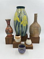 Front view showing all six pottery items on wood blocks including large iris vase, tall beige vase, small striped pot, two small marked pots, and purple pot