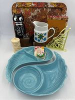 Wide shot showing all items in the lot laid out on a white surface including the mid-century turquoise pottery bowls, black cat clock, floral metal tray, white pitcher, trivet, and other decorative items.