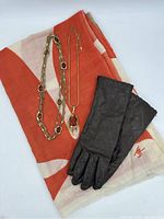Photo shows two gold-tone necklaces, one with oval amber stones and one pendant style, lying on orange and cream geometric wool throw with JR embroidered logo, and a pair of vintage black leather lined gloves.