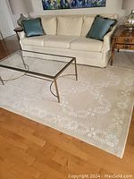 Full view of cream and beige area rug placed under a glass coffee table and in front of a cream sofa, showing floral leaf border and diamond lattice central pattern