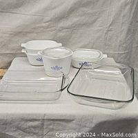 Overall shot of all Corningware casserole dishes and glass bakeware pieces on white fabric background with soda can for scale.