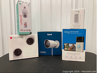 Front view of all cameras in original boxes including Ring Video Doorbell, Ring Chime Pro, Kami Outdoor Security Camera, Yi Home Camera 3 Family Pack 2-in-1 and a WiFi Smart Camera bulb device.