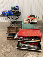 Full view of the lot showing red Craftsman two-drawer toolbox open with hand tools inside, blue plastic storage bins on a folding table, power tools in cases, and assorted boxes stacked nearby.