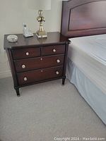 Full view of one nightstand beside bed with lamp, showing two small top drawers and two larger bottom drawers, silver tone knob handles, dark mahogany finish, rounded tapered legs.