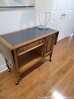 Full view of the tea cart showing oak finish, black tabletop, drawer, open shelf, and cabinet door along with caster wheels.