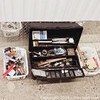 Photo of open black plastic tool box showing multiple compartments filled with various hand tools and small hardware items. Two white plastic baskets flanking tool box also contain assortments of hand tools and accessories.