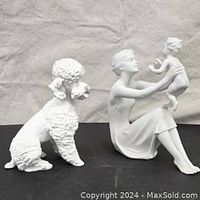 Front view of white porcelain figurines: woman holding child and sitting poodle dog.